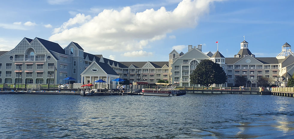 A wide waterfront view of Disney’s Yacht Club Resort at Walt Disney World, featuring a large grey-and-white New England–style hotel with gabled roofs and balconies. Several boats are docked along the marina in the foreground, with blue and red umbrellas lining the pier. Calm water stretches across the bottom of the image under a bright sky with fluffy white clouds.