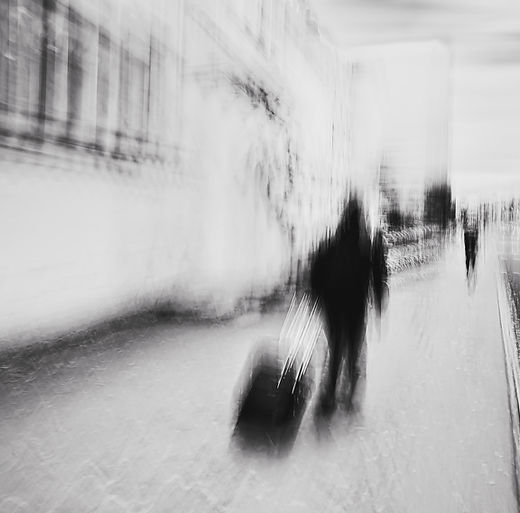 A contemporary black-and-white abstract photograph capturing the fleeting motion of a traveler with a suitcase. The blurred figure—a wandering soul—evokes mystery and solitude, symbolising the transience of urban life and the continuous rhythm of human journeys. A Limited Edition Digital Graphic Artwork by Douglas Carr.