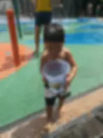 Child holding a bucket at a colorful splash pad, surrounded by fountains. An adult stands nearby. It's sunny, and the mood is playful.