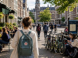 American student walking through a European university city with backpack, cyclists, cafés, and historic buildings