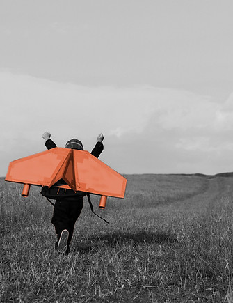 A little boy with hands up in the air running across a field with an orange cardboard jet attached to his back