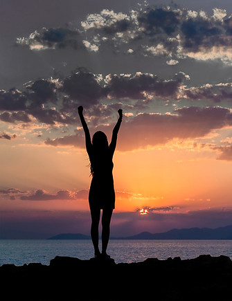 A woman standing on sand over looking the water and sunset with her hands up in the air