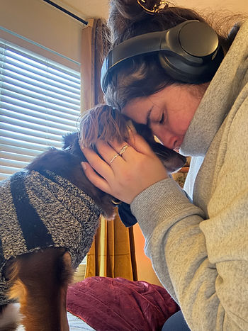 A woman wearing headphones lovingly embraces a dog in a sweater. The indoor setting has soft lighting and a window with blinds.