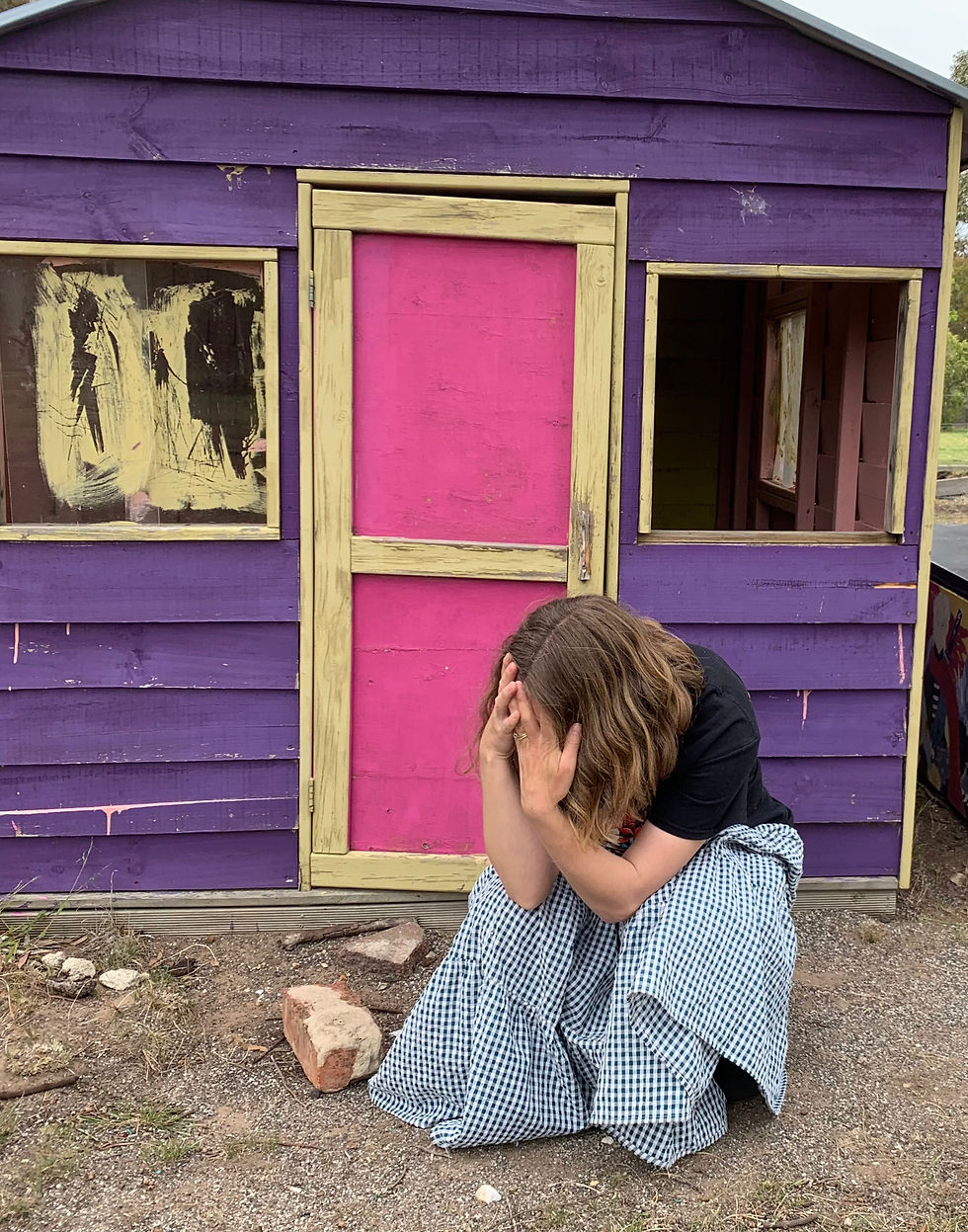 Person crouches, hands covering face, in front of a purple playhouse with a pink door. Gingham skirt, outdoor setting, somber mood.