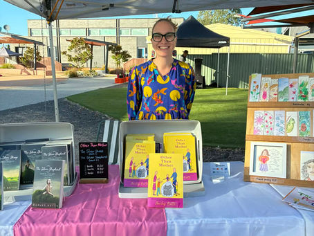 Smiling person stands under a tent with books displayed on a table. The table has a colorful banner. The background shows buildings.
