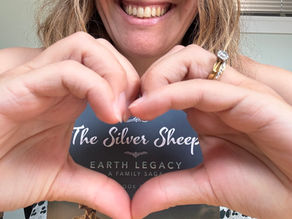 Smiling woman in glasses makes a heart with her hands while holding The Silver Sheep book; cozy indoor background.