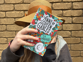 A girl wearing a straw hat holds a colourful copy of the book 'Anne of Green Gables' in front of her face