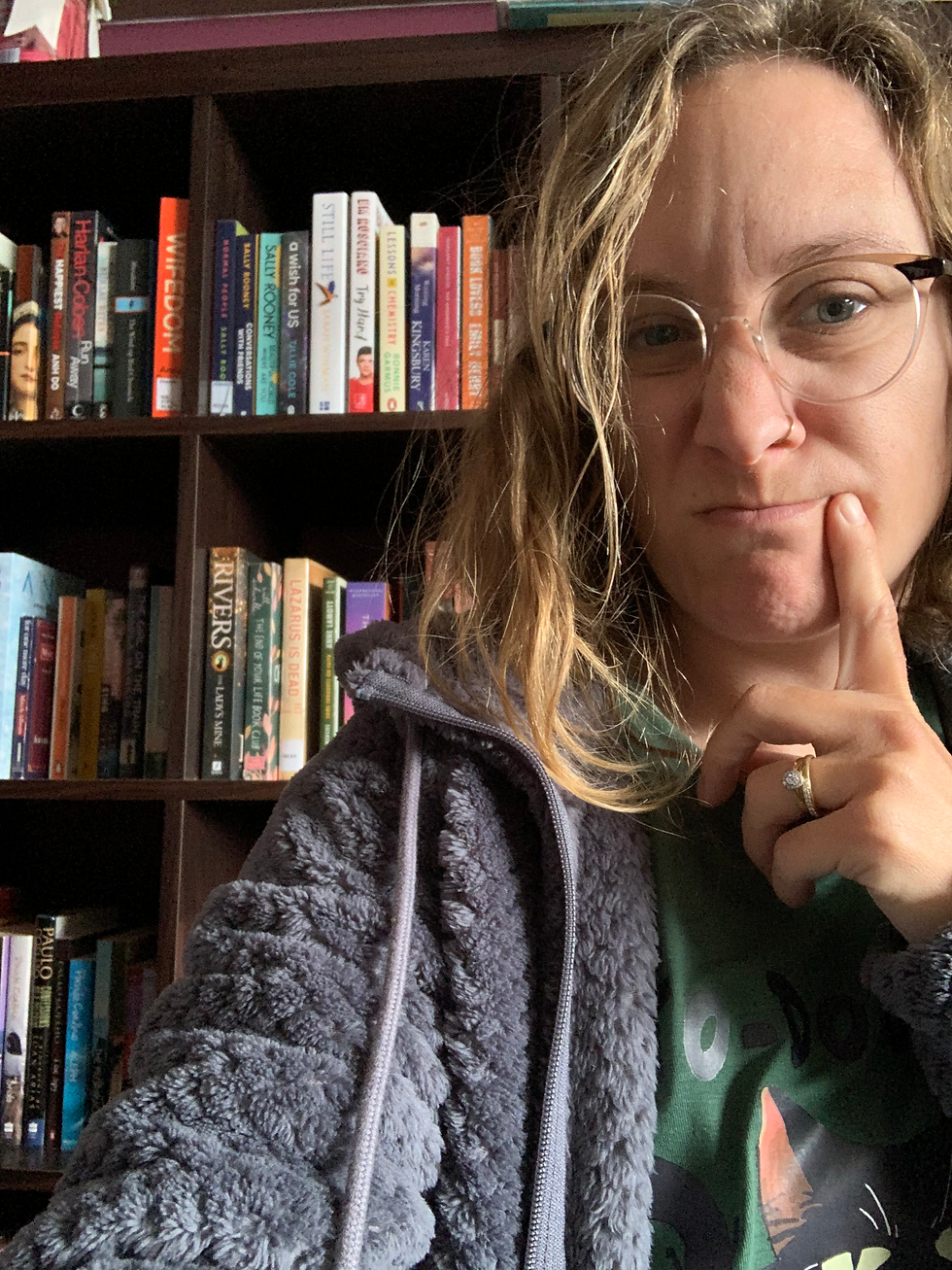 Woman with glasses posing pensively, wearing a gray fleece. Bookshelf in the background with visible book titles. Cozy indoor setting.