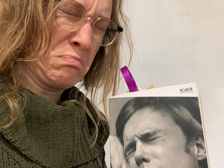 Woman frowning while holding the book "A Little Life" by Hanya Yanagihara. The cover shows a person in distress. She wears glasses and a green sweater.
