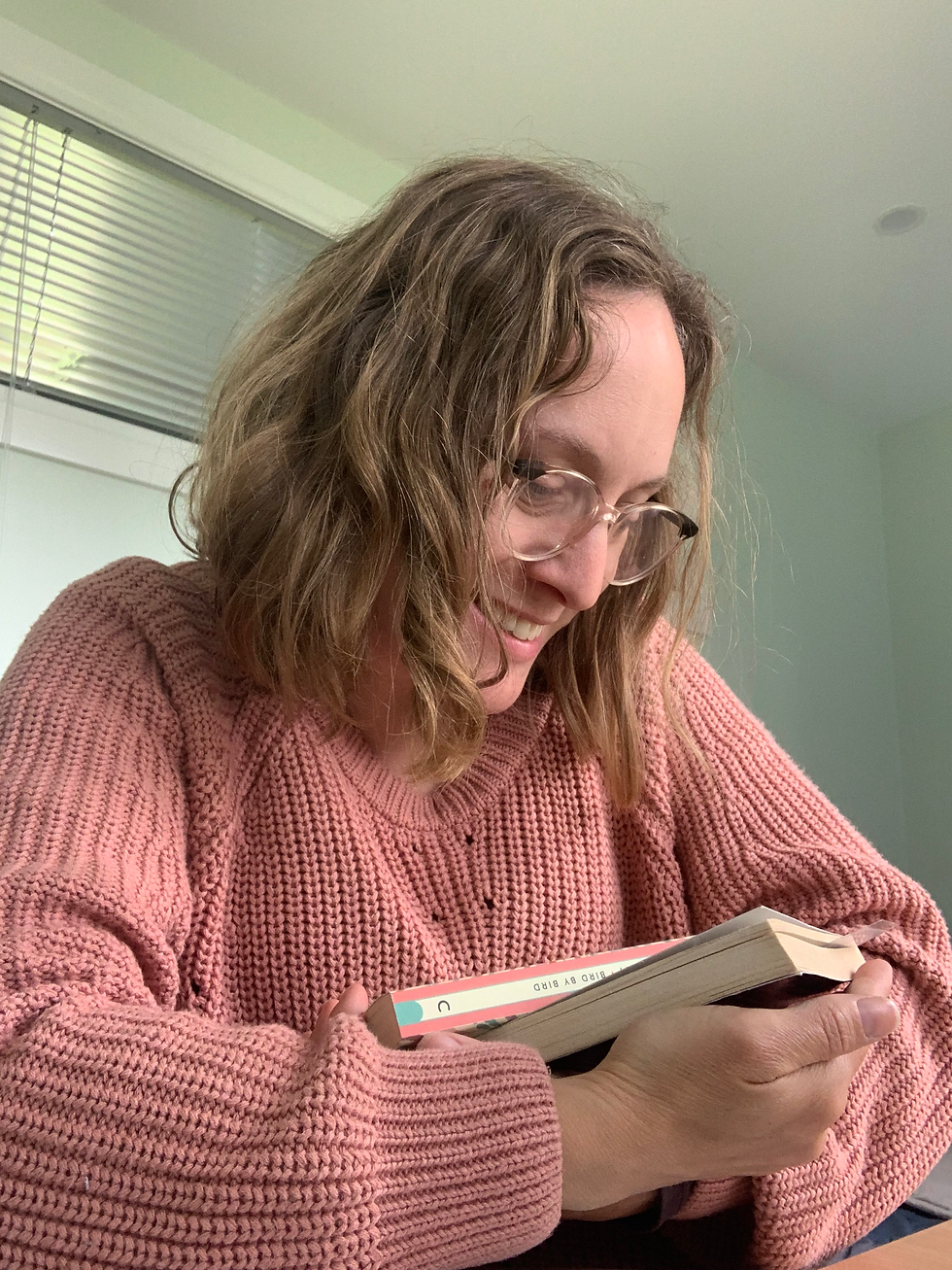 Woman in a pink sweater smiles while cradling some books in her arms. Soft lighting creates a cozy, relaxed atmosphere.