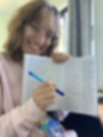 Smiling person with glasses holds a notebook displaying handwritten notes, pointing with a blue pen. They're in a brightly lit room.