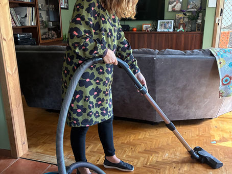 Person in a floral dress vacuums wooden floor in a cozy living room with records on the wall. Mood is calm and domestic.
