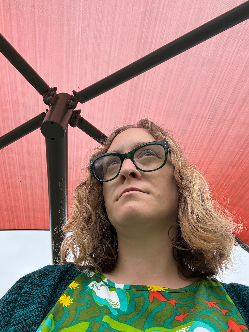 Woman with glasses and wavy hair under a pink canopy, wearing a colorful floral top and looking upward thoughtfully.