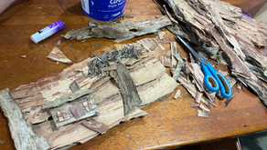A picture of a house and tree made from papebark lies on a table. Scissors, glue, bark, and a yoghurt bucket filled with sticks and leaves surround the bark picture.