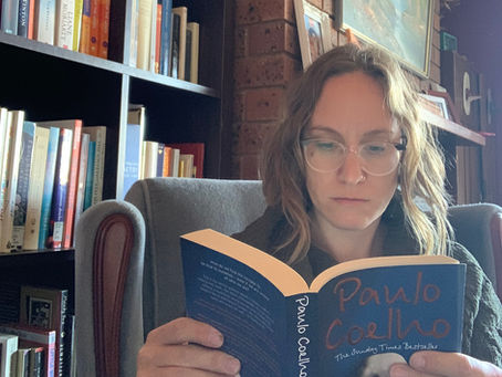 A person with glasses reads "Paulo Coelho" in a cozy library with a brick wall and bookshelves. Calm and focused atmosphere.