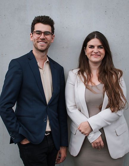 Zwei Menschen stehen nebeneinander. Links ein großer Mann in blauem Anzug. Rechts eine Frau in beigem Jacket und langer Haare