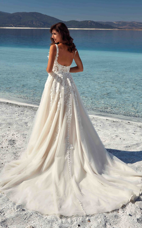 Eddy K wedding dress with beaded lace with sequins, 3D appliqués, tulle, unlined bodice, slit, buttons to end of train on woman standing by crystal blue water.