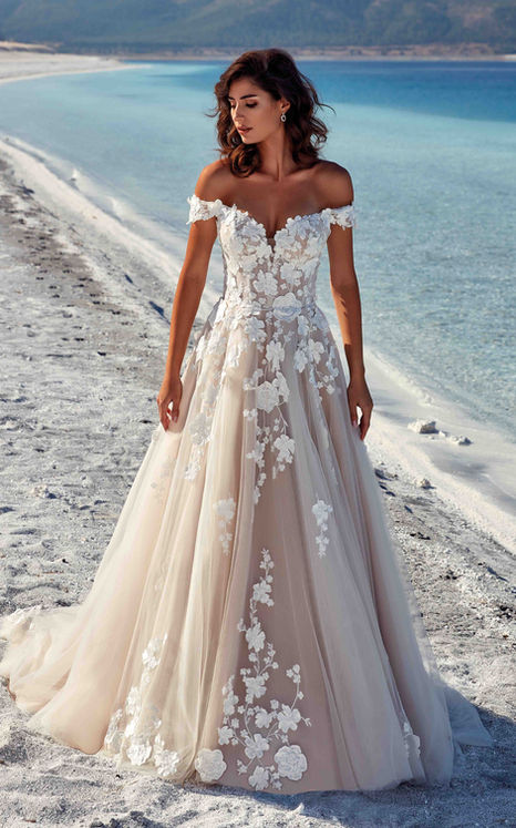 Eddy K ballgown with ivory lace over champagne tulle, 3D appliqués and mocha matte lining and buttons to end of train. Standing on the beach by blue water.