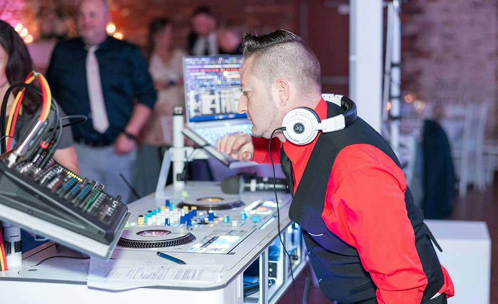 DJ wearing headphones at a mixing console with colorful controls, set in a dimly lit event room. A blurred crowd is visible in the background.