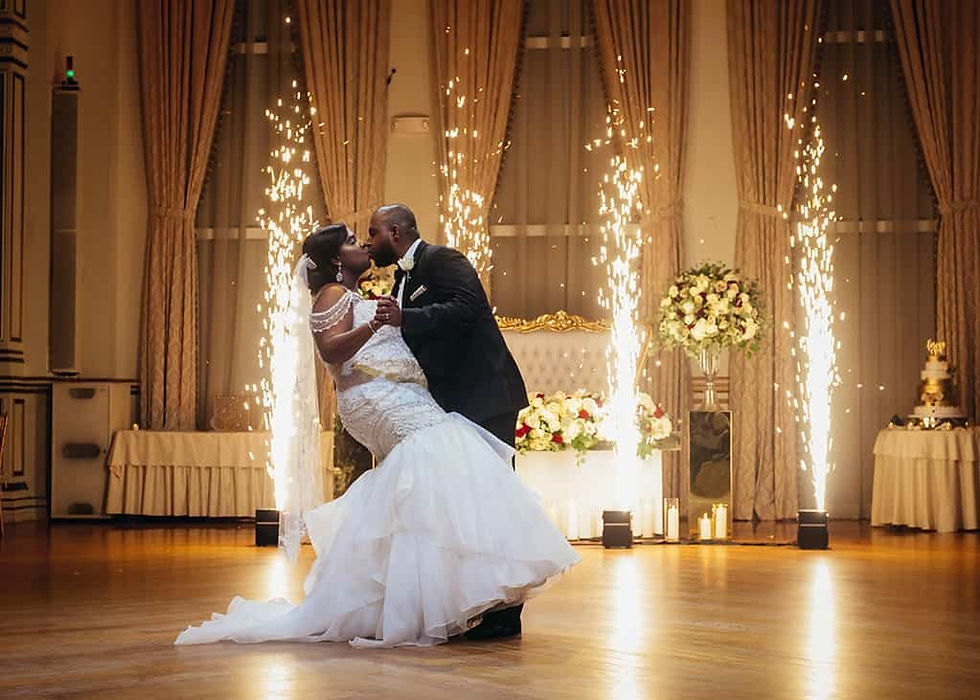 A couple shares a romantic dance in a grand hall. Fireworks light up the background with floral decor, creating a celebratory atmosphere.