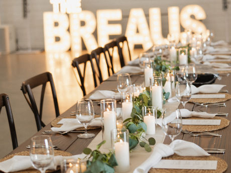 Candlelit banquet table set for a wedding, with greenery, white napkins, glassware and glowing THE BREAUS letters in back.