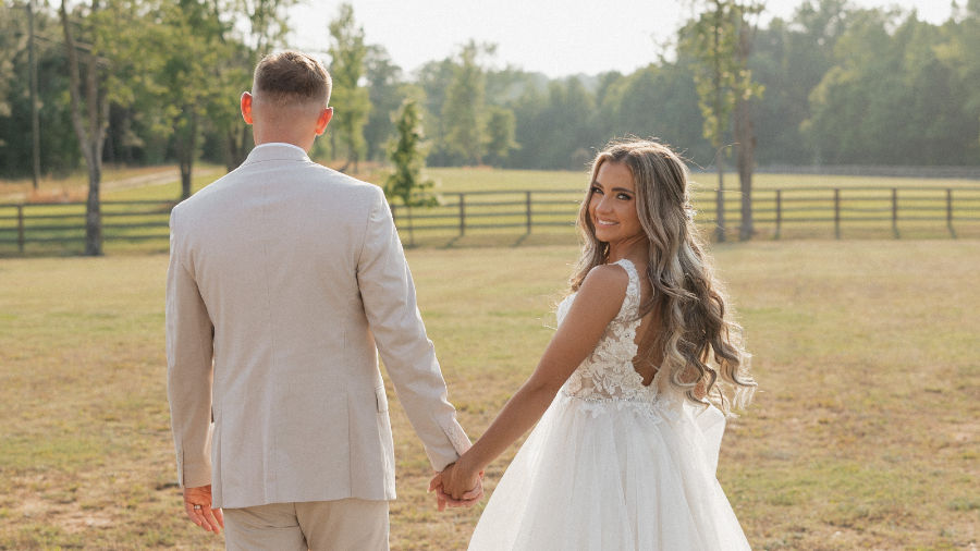 Bride in white wedding dress looks back smiling while holding hands with groom in beige suit in a sunny field with fence and trees