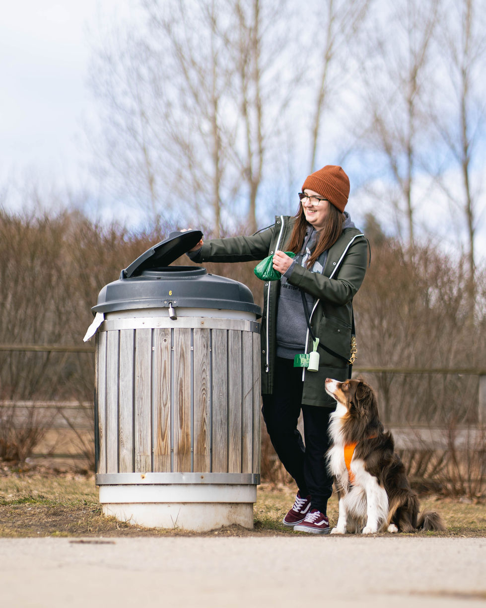 Woman with long brown hair opening a large garbage can while her Red Tri Australian Shepherd stands behind her as she puts a green Earth Rated poop bag in the garbage can.