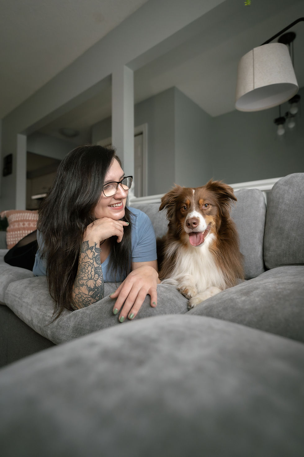 Smiling woman lying on a gray couch beside a happy brown tri-color Australian Shepherd, both resting on a plush Nolan Interior washable couch cover.