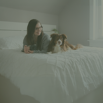 Canadian pet and lifestyle content creator Carly from Her Cozy Crew holding a camera on a white bed beside her Australian Shepherd dog, offering brand photography and UGC services