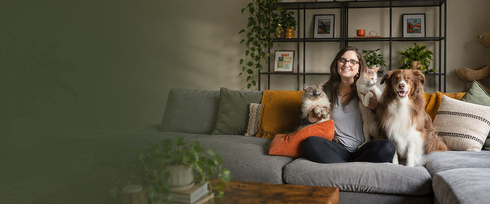 Carly sits on a gray couch with her two cats and Australian Shepherd in a cozy, plant-filled living room