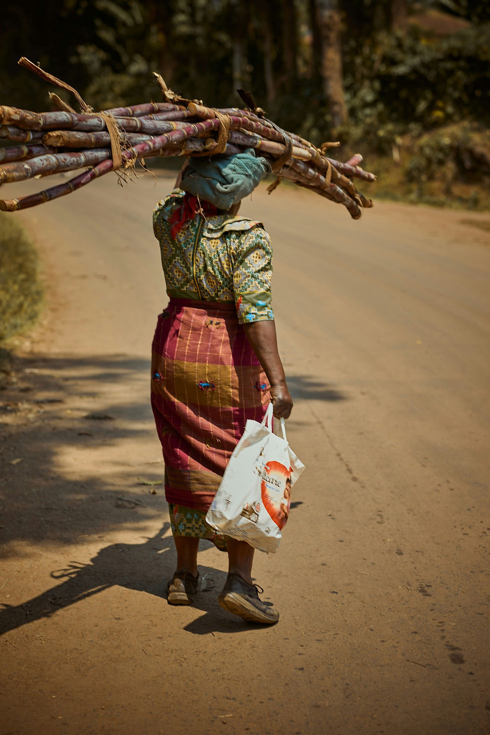 A woman in colorful attire carries sugarcane on her head and a bag on a rural road. Sunlight casts shadows, with greenery in the background.