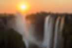 Sunset over Victoria Falls with cascading water, mist rising, and an orange sky. Cliffs frame the waterfall, creating a serene scene.