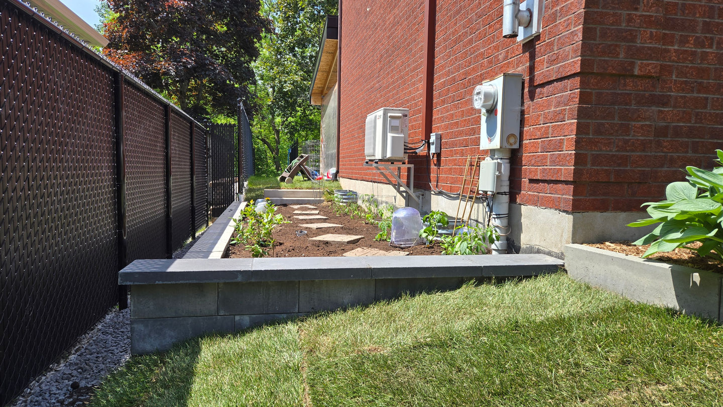 Jardin potager longeant la maison en brique rouge, bordé par un muret en béton gris