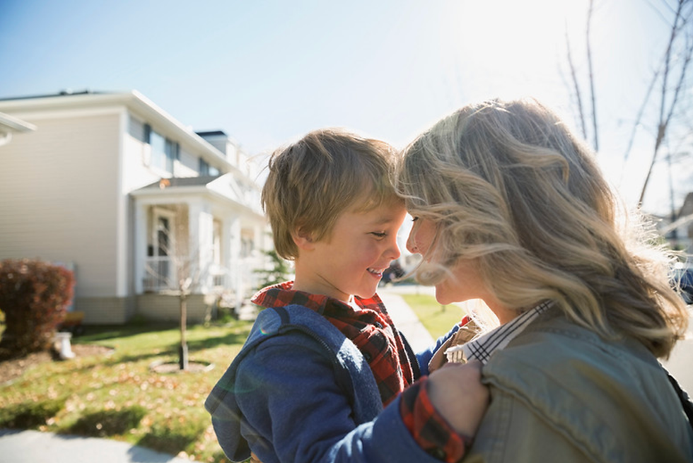 A smiling child and woman embrace outside a house on a sunny day. The child wears a blue jacket, and the mood is joyful and warm.