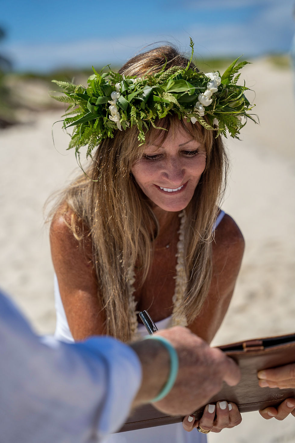 Thumbnail: Smiling bride in white with floral accessories stands on beach, holding bouquet in celebration