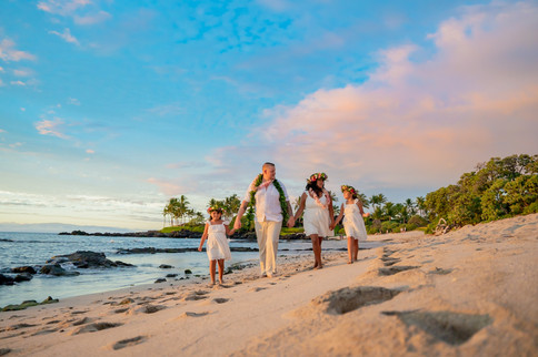 Group in white walks hand-in-hand at Kukiʻo Beach sunset, framed by palms, ocean, and pastel sky.