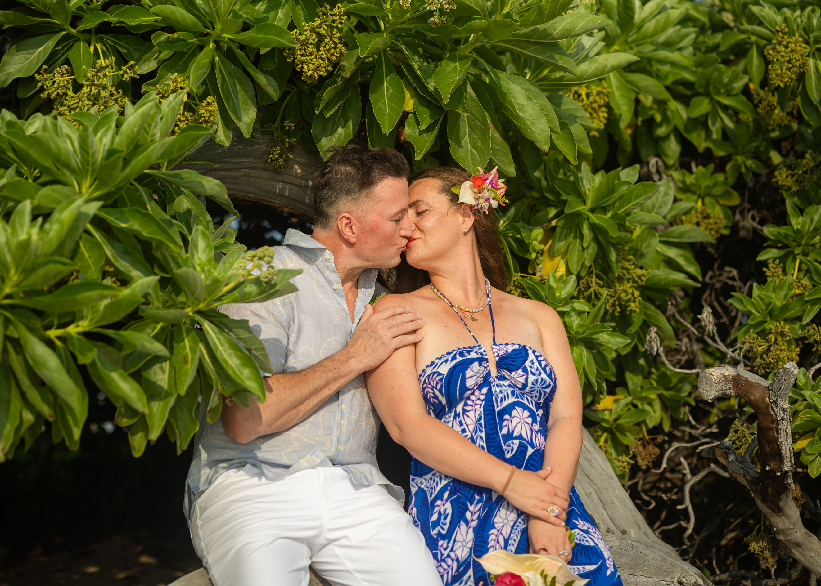 Couple in pink leis celebrates on Kona beach at sunset, arms outstretched in tropical joy.
