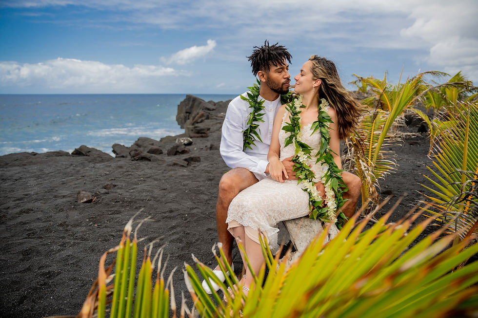 Ginelle and Tariq - seated on a small bench at Kaimu Black Sand Beach, Kalapana, Hawaii, during the post-wedding elopement photography session. Couple adorned in traditional Hawaiian wedding leis.