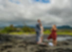 Couple stands barefoot on rocky shore in front of officiant blowing conch shell. Overcast sky and green landscape in background.