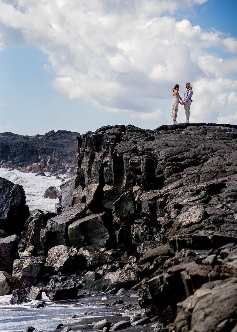 Oxana and Max stand amongst the tall volcanic cliff, hand in hand, just married at Kaimu Black Sand Beach, Hawaii, with Kona Wedding Officiant.