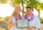 Two people smiling, wearing purple leis, holding a "Married in Hawaii" certificate on a sunny beach with trees and a bright background.