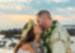 A couple smiling and touching noses adorned with flower leis and garlands, against a serene rocky Big Island Hawaii beach and sunset sky backdrop.