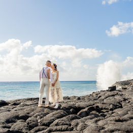 Oxana and Max kiss along the sweeping volcanic cliffs of Hawaii's Kaimu Black Sand Beach - Pacific waves crashing along the rocks behind them. 
