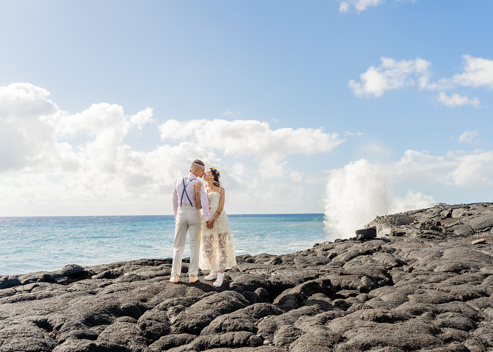 Oxana and Max kiss along the sweeping volcanic cliffs of Hawaii's Kaimu Black Sand Beach - Pacific waves crashing along the rocks behind them.