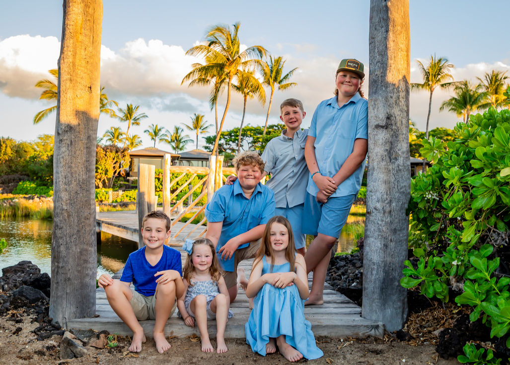 Group of six kids posing under wooden dock with palm trees, blue sky, and tropical coastal background