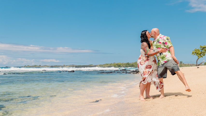 Couple kissing on a sandy beach in Kailua Kona, Hawaii, with blue skies, ocean waves, and tropical trees in the background.