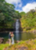 Hawaii wedding couple embracing near a waterfall with lush greenery and a bright blue sky. The woman wears a floral crown, creating a joyful, romantic vibe. Married with Kona Wedding Officiant.