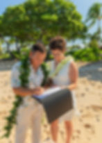 A couple signs Hawaii marriage license on Kuki`o beach. Both wear white and Hawaiian leis. Green trees and blue sky in the background.