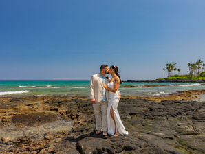 Bride and groom kissing on a lava rock outcropping with turquoise ocean and palm-covered point in the background, Big Island Hawaii wedding.