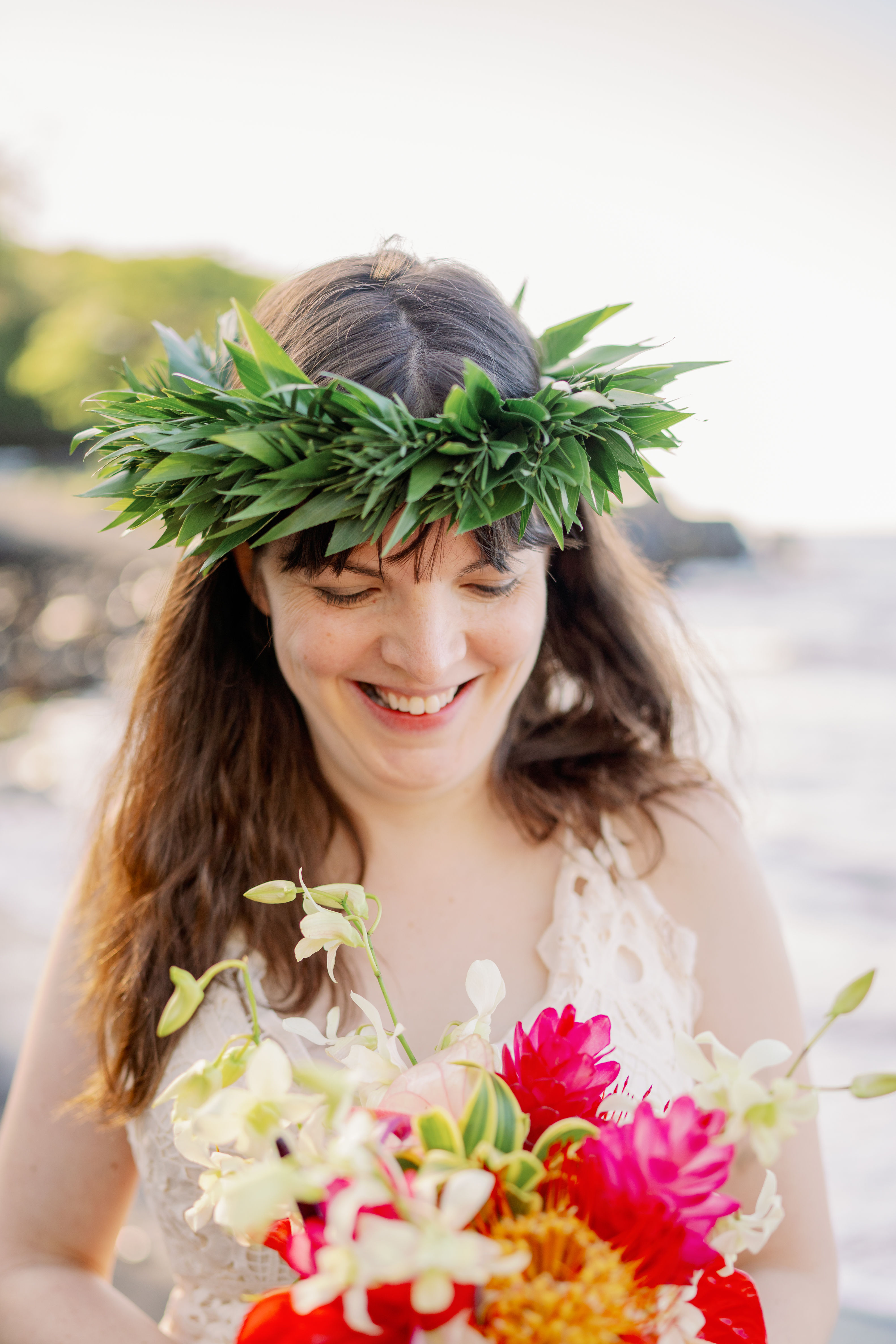 Person in white with leafy crown and bouquet stands near ocean, framed by cliffs, water, and greenery